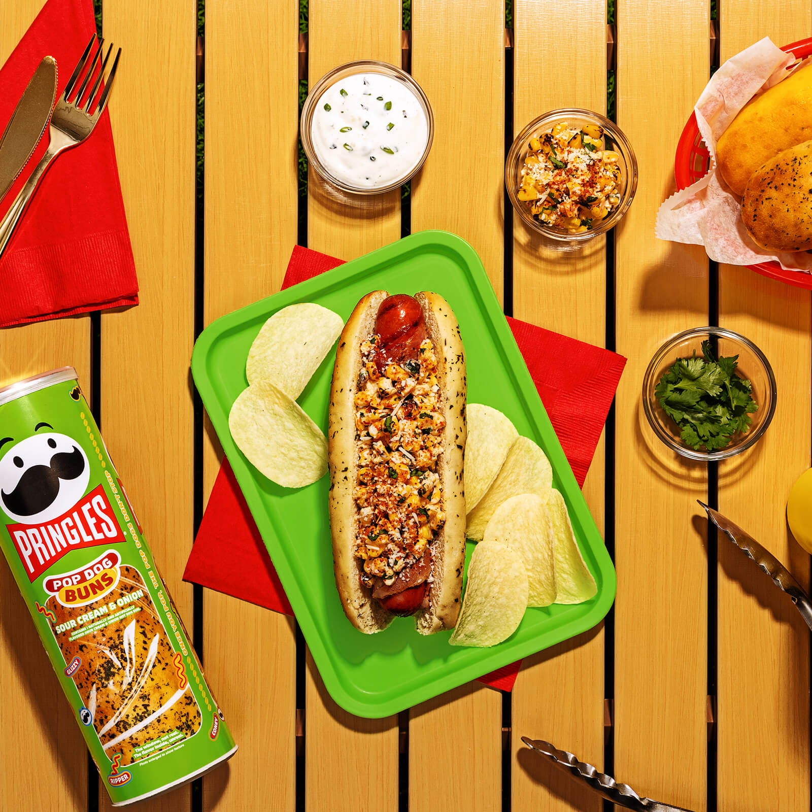 A bacon-wrapped hot dog in a Pringles Sour Cream & Onion bun, topped with esquites, cotija cheese, and chili lime seasoning, served on a green tray with Pringles potato crisps.