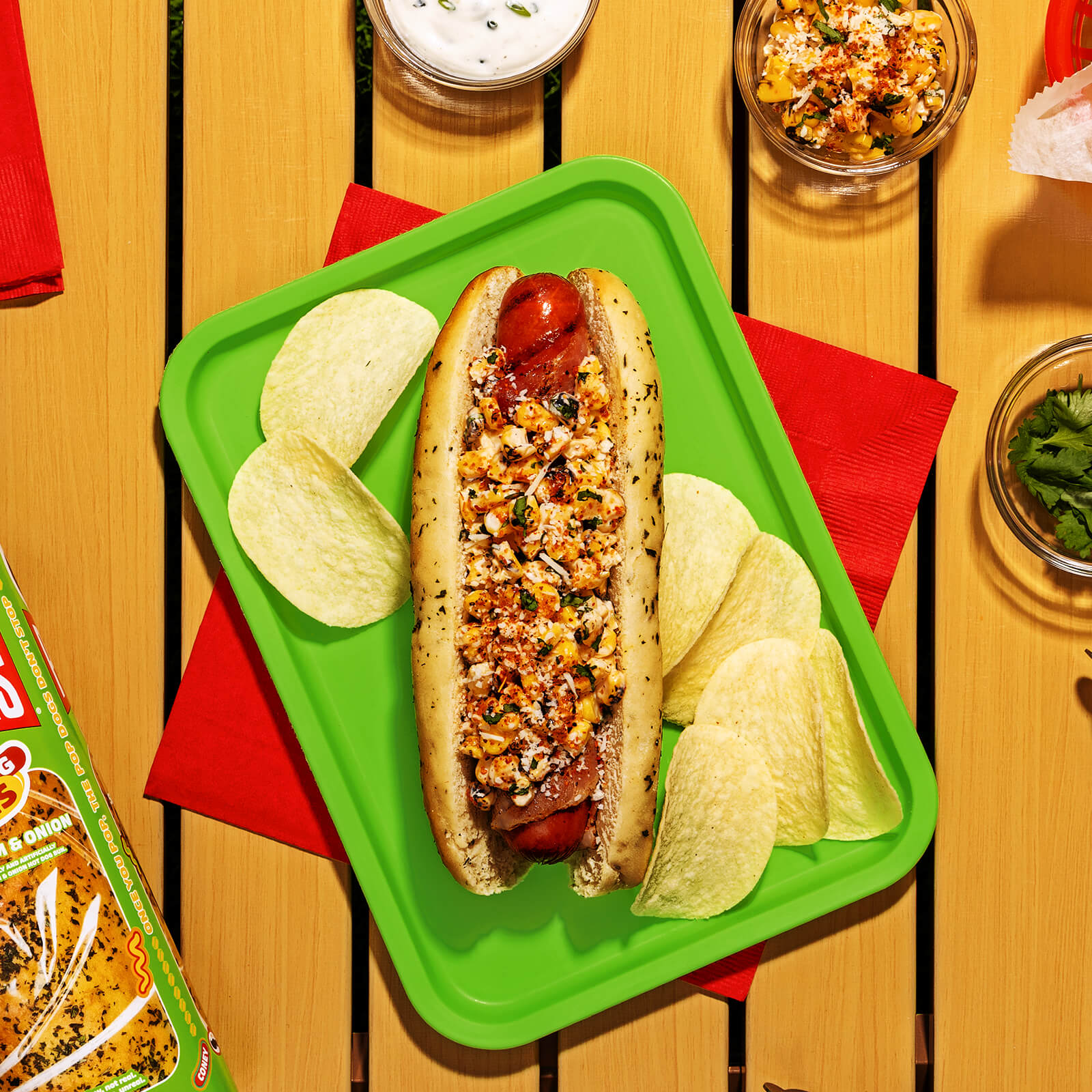 A bacon-wrapped hot dog in a Pringles Sour Cream & Onion bun, topped with esquites, cotija cheese, and chili lime seasoning, served on a green tray with Pringles potato crisps.