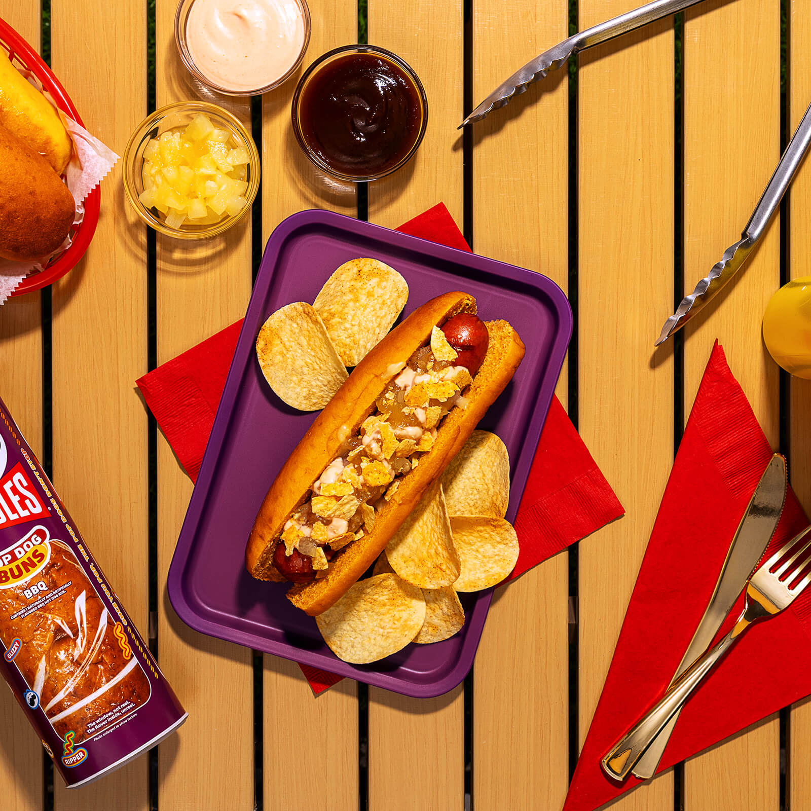 A cooked hot dog in a Pringles BBQ bun, topped with melted mozzarella, pineapple jam, pink sauce, and crushed Pringles BBQ potato crisps, served on a purple tray.