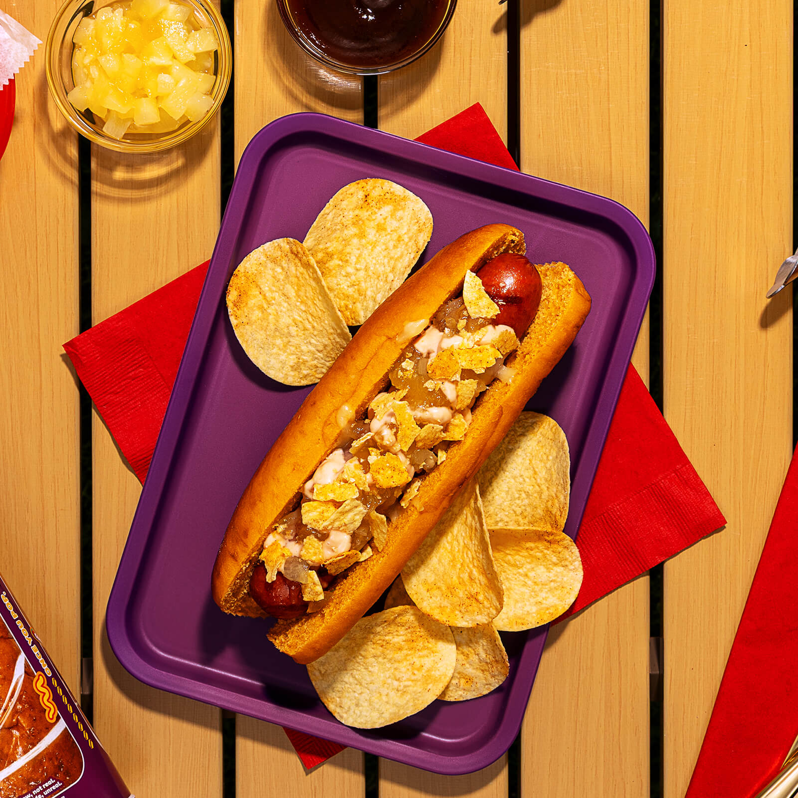 A cooked hot dog in a Pringles BBQ bun, topped with melted mozzarella, pineapple jam, pink sauce, and crushed Pringles BBQ potato crisps, served on a purple tray.
