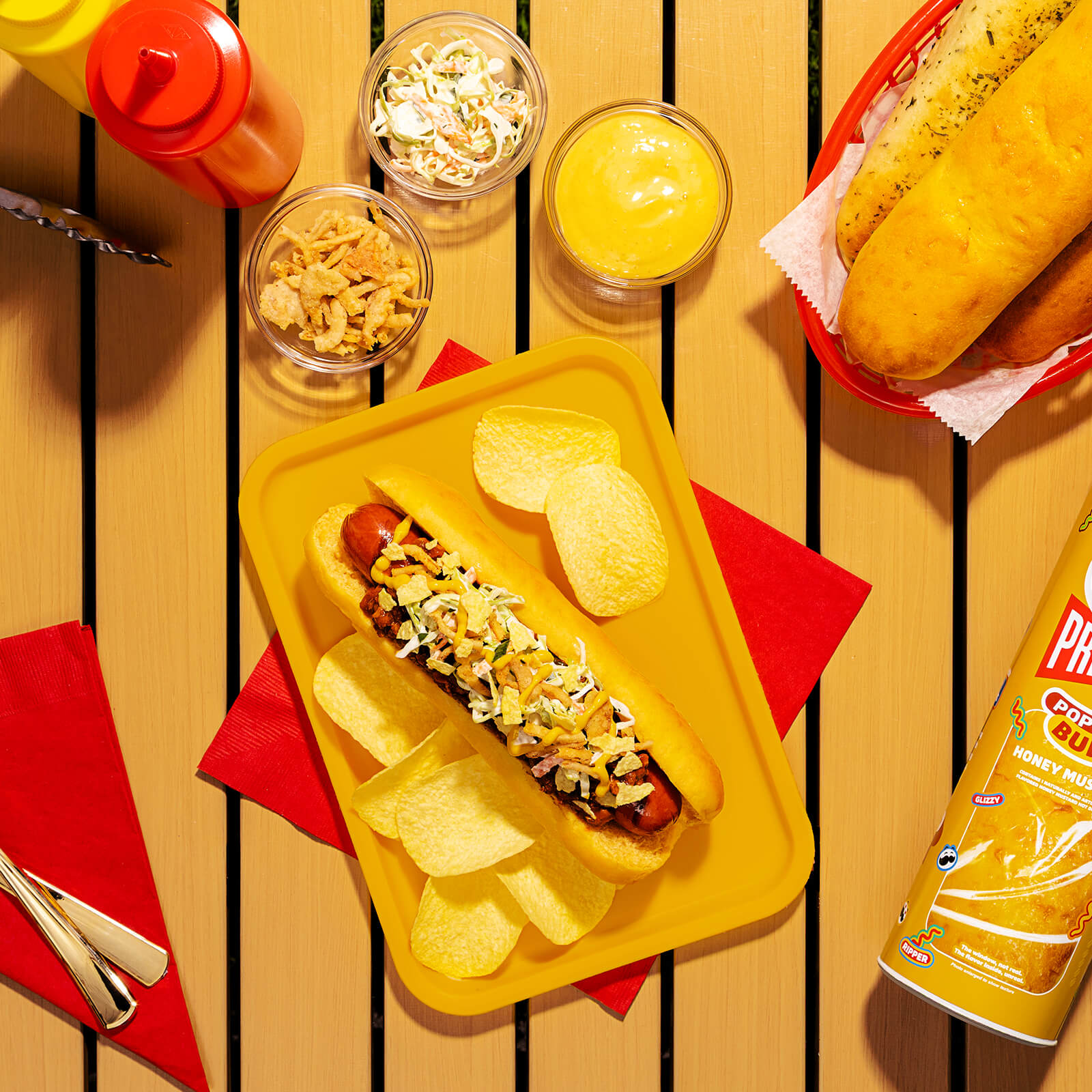 A cooked hot dog in a Pringles Honey Mustard bun, topped with chili, coleslaw, crispy onion strips, mustard, and crushed Pringles Honey Mustard potato crisps, served on a yellow tray.