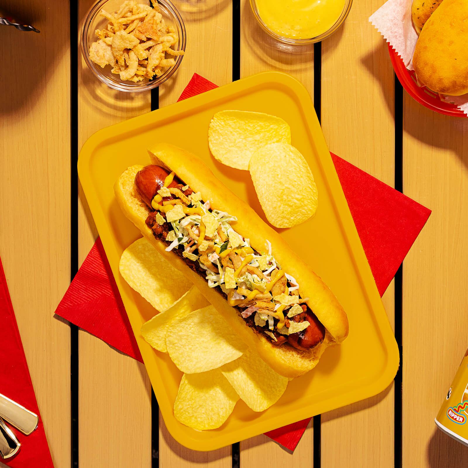 A cooked hot dog in a Pringles Honey Mustard bun, topped with chili, coleslaw, crispy onion strips, mustard, and crushed Pringles Honey Mustard potato crisps, served on a yellow tray.