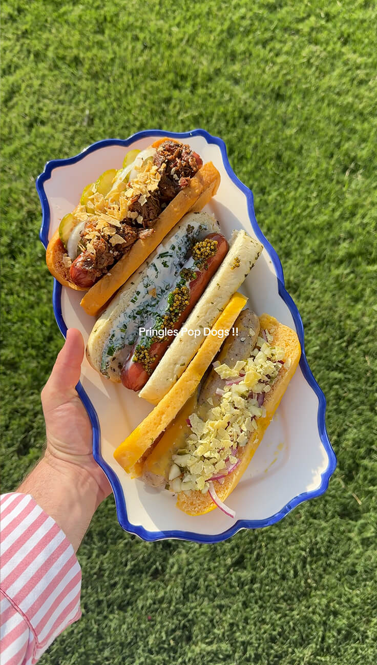 Video thumbnail, A hand holds a platter of three topped Pringles Pop Dog Buns hot dogs over grass.